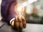 A hand holding a lit lightbulb next to a laptop computer.