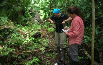 Photo of a scientist collecting a soil sample using an auger and another scientist taking notes.