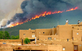 Photo of a wildfire over pine forested hills in background with houses in foreground in New Mexico.