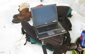 Photo of a laptop downloading data, backpack, hiking boots and instruments in the snow.