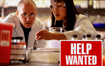two scientists with a Help Wanted sign in the foreground.