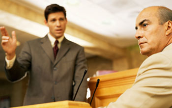 a seated witness and a standing lawyer with a raised hand in a courtroom.