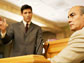 Image of a seated witness and a standing lawyer with a raised hand in a courtroom.