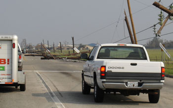 Photo of vehicles traveling down a road with downed telephone poles and collapsed houses.