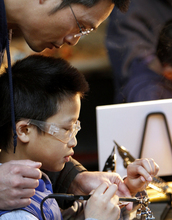 A father and son work on soldering to make an automaton out of copper wire.