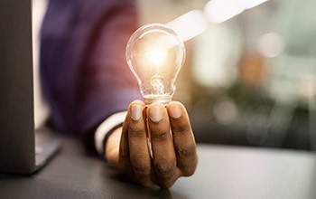 A hand holding a lit lightbulb next to a laptop computer.