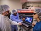 Three people wearing lab coats and goggles working in a lab with computer data.