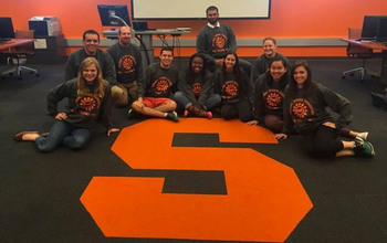 A group of smiling students pose around a large S on the floor of a classroom at Syracuse University.