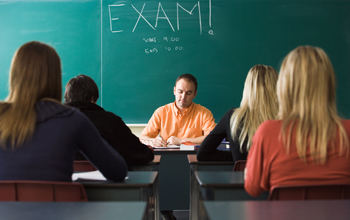 Photo of a teacher sitting at a desk , blackboard with the word EXAM, and students taking a test.