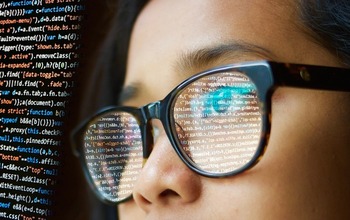 Close of a woman wearing glasses. Computer code is reflected in the lenses.